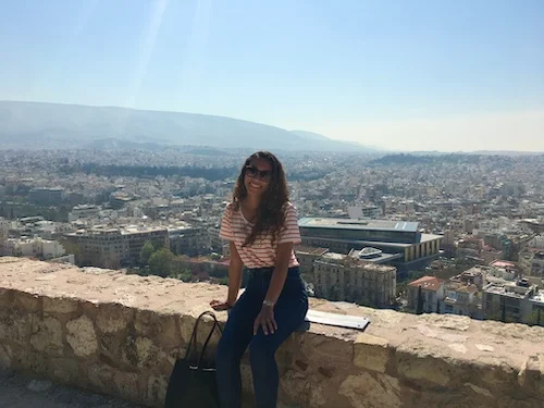 Journalist Jenzia Burgos sitting on ancient stone walls at the Acropolis in Athens, Greece as a study abroad student in 2017, overlooking the city below the Parthenon.