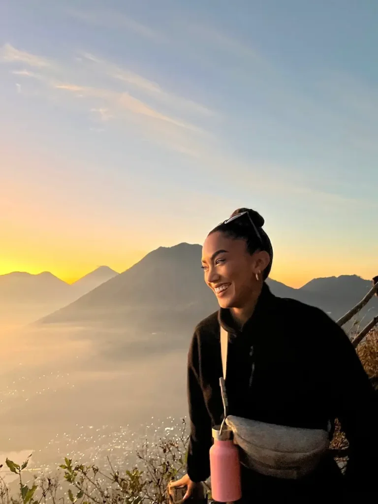 Journalist and travel creator Jenzia Burgos smiling at sunrise on the Rostro Maya hike (Indian Nose) in Guatemala, with volcanic mountain ranges and Lake Atitlán visible in the misty background.