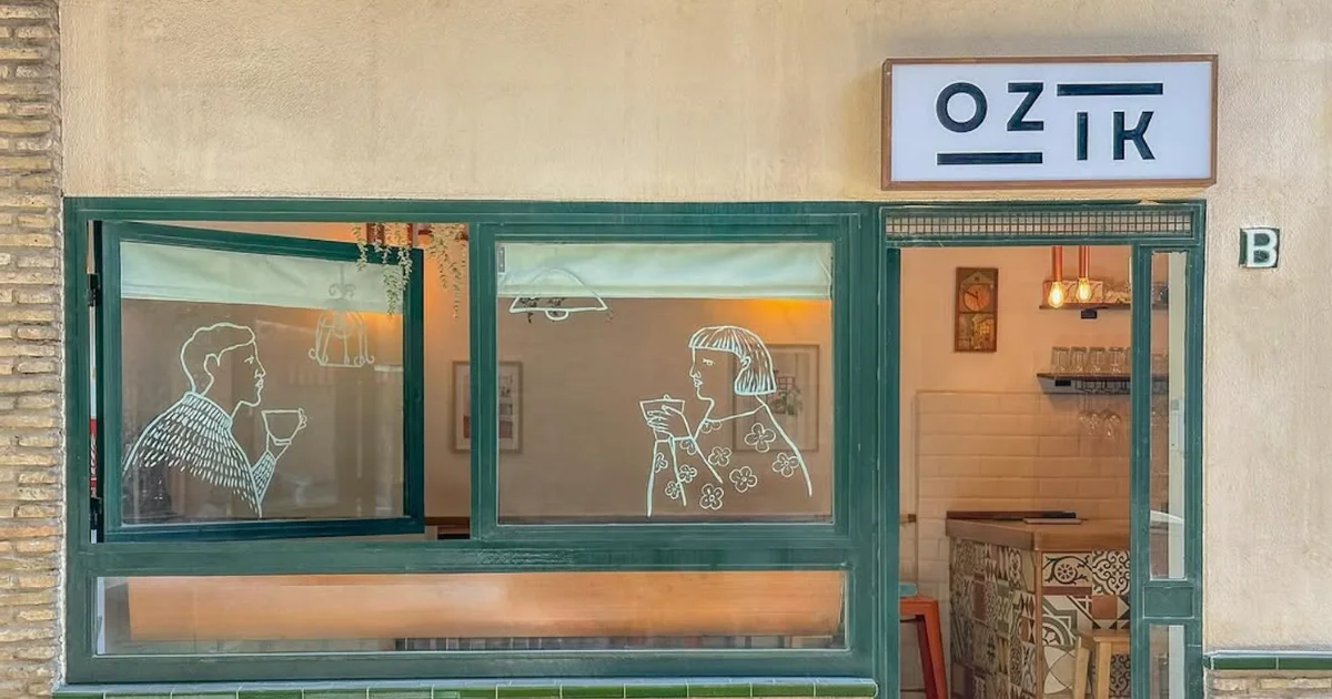 Facade of Ozik café with green-framed windows decorated with hand-drawn illustrations of two people drinking coffee, exposed brick detailing, and a clean white logo sign above the entrance.