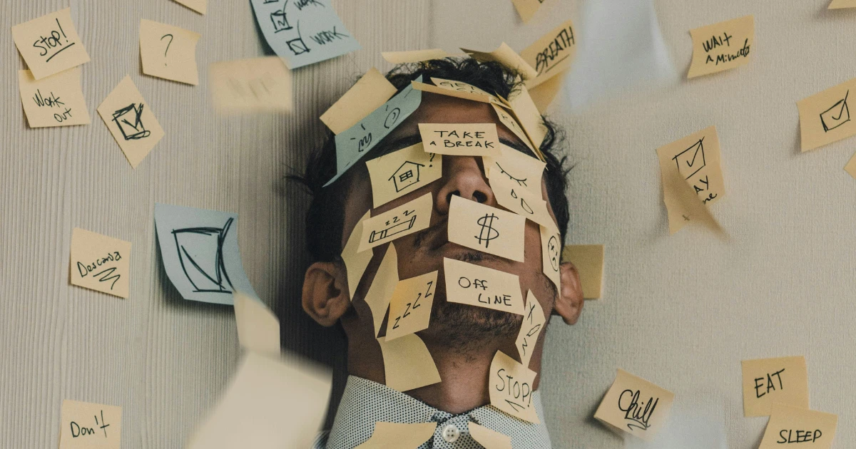 A person lying back with sticky notes covering their face and surrounding them, notes reading "Take a Break," "Work," "Stop," and "Off Line," representing the overwhelming paperwork process of applying for NALCAP