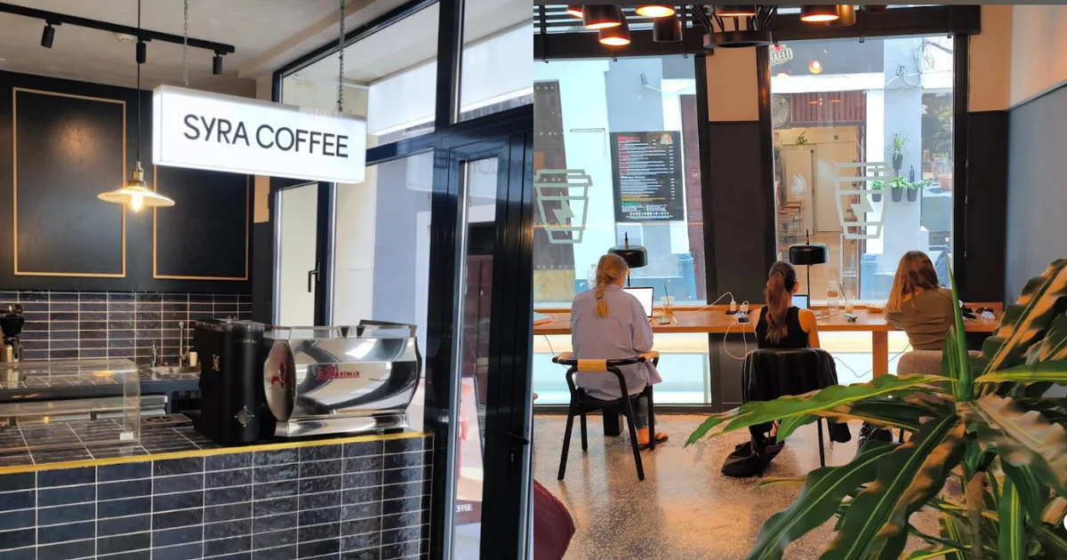 Three people working on laptops at a laptop-friendly café in Seville, Spain, seated at a communal window bench; left side shows a dark tiled bar with gold accents and an illuminated Syra Coffee sign.