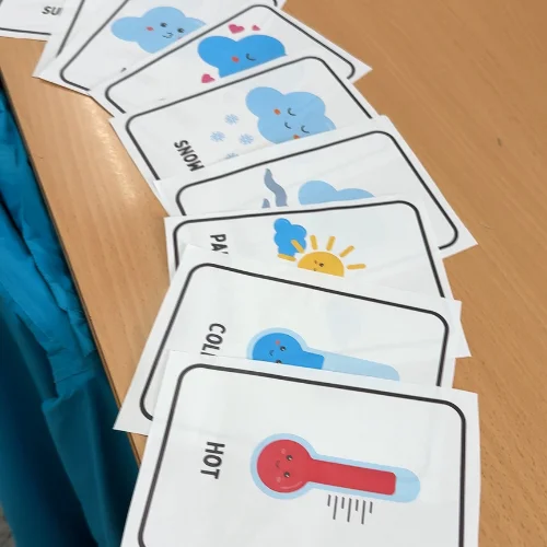 Weather vocabulary flashcards laid out on a classroom desk, including hot, cold, sunny, and snow, used to teach English in Spain
