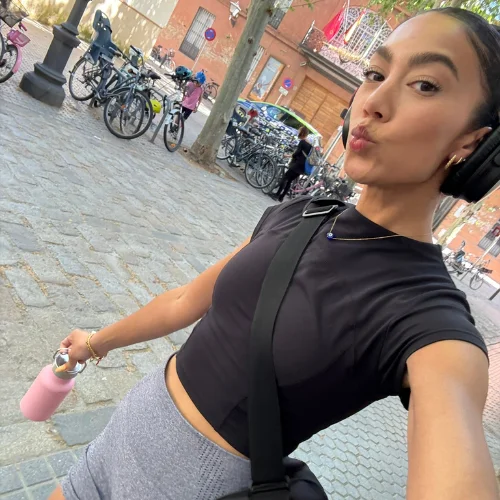 Woman taking a selfie on a cobblestone street in Spain after a workout, wearing headphones and holding a water bottle