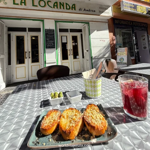 Outdoor table at La Locanda di Andrea restaurant in Sevilla, Spain, with grilled bread topped with garlic and herbs, olives, and a glass of tinto de verano.