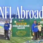 Collage of travel blogger and journalist Jenzia Burgos at the 2026 NFL Melbourne Game between the LA Rams and San Francisco 49ers, posing in Rams gear in front of "2026 NFL Melbourne Game" signage, with the packed stadium and football field in the background, text overlay reads "NFL Abroad."
