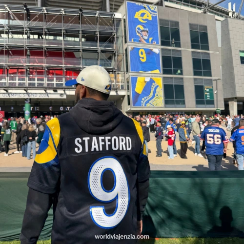 A fan in a Matthew Stafford No. 9 LA Rams jersey stands with his back to the camera outside the stadium entrance, with a large Rams banner on the building ahead.