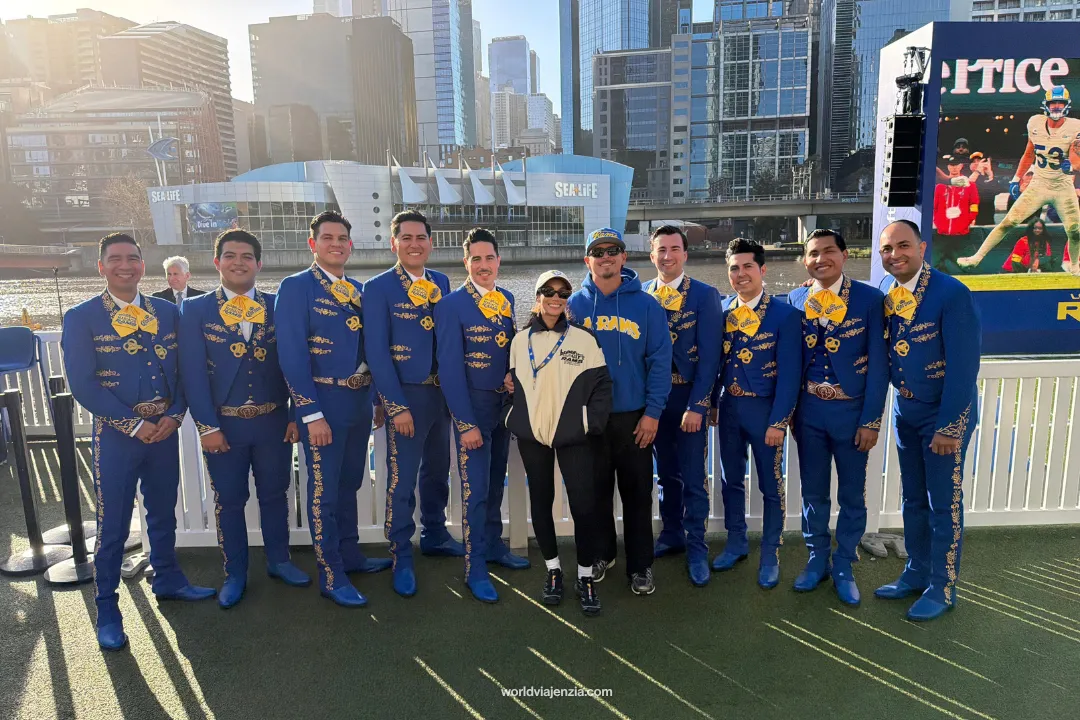 Travel journalist Jenzia Burgos and her partner pose with a mariachi band in royal blue embroidered suits along the Yarra River at the NFL Melbourne fan fest, with the city skyline in the background.