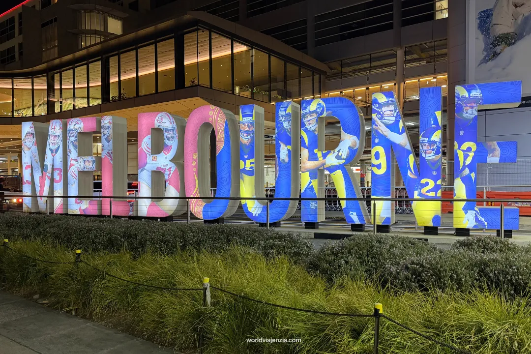 The illuminated MELBOURNE sign outside Melbourne Airport at night, with NFL player imagery projected across the letters in 49ers red and Rams blue and gold.